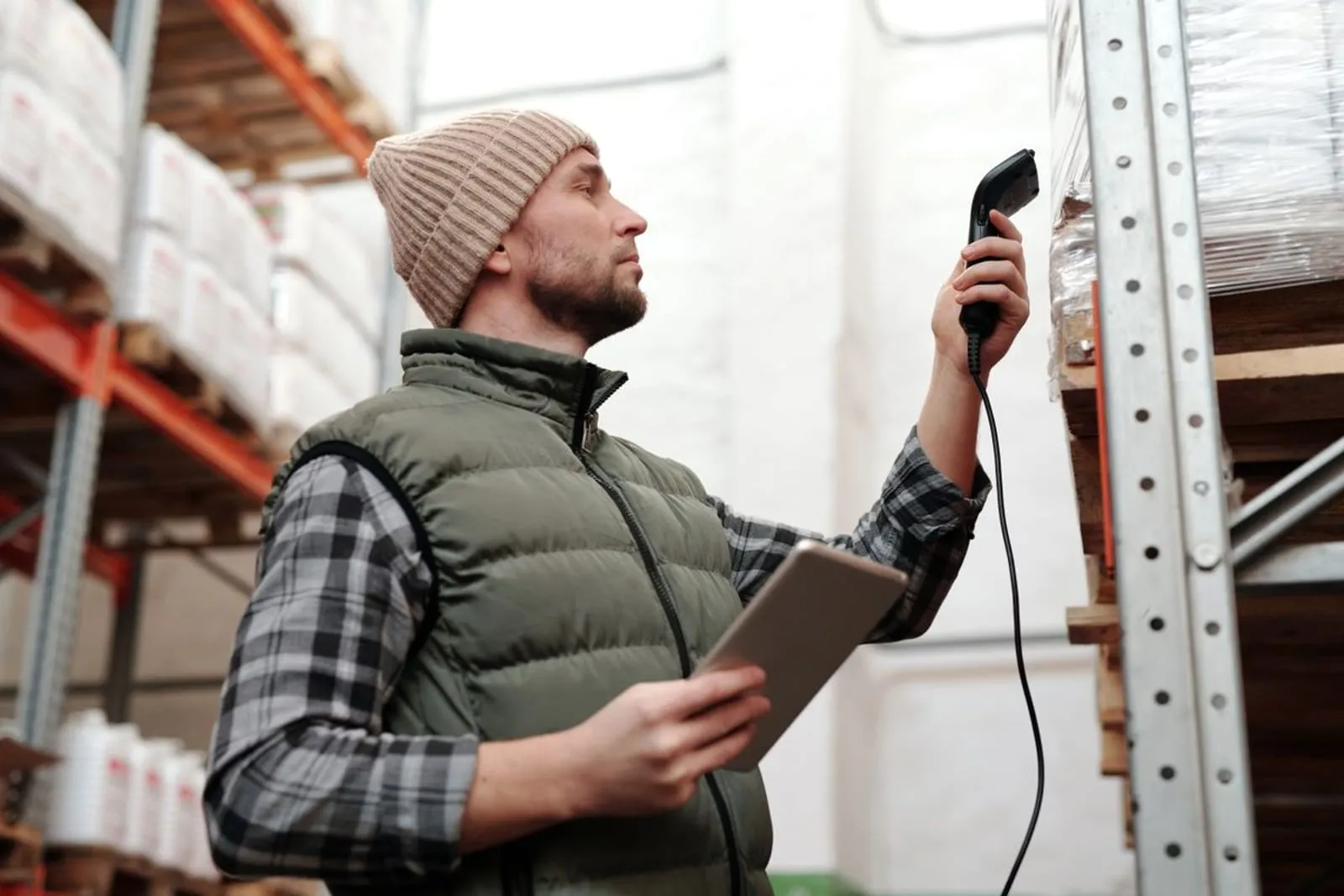 A warehouse worker holding a handheld scanner in one hand and a tablet in the other, checking stock on a rack